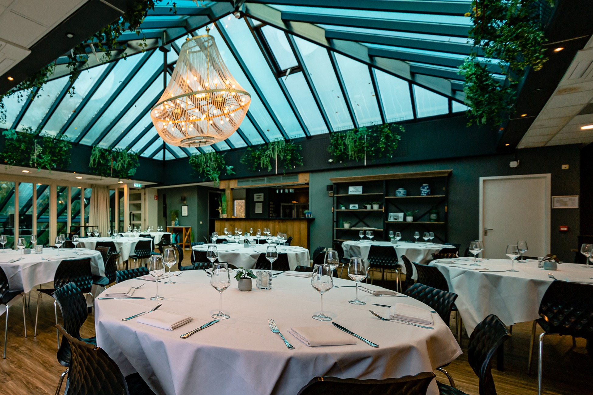 De Beatrixpark zaal in Amstel Boathouse, groot dakraam en bar, planten en groen, ronde tafels, diner opstelling